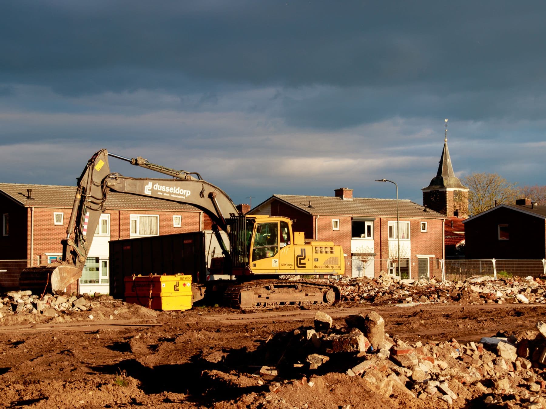 A yellow excavator on a cleared lot of red soil, with brick houses behind it.