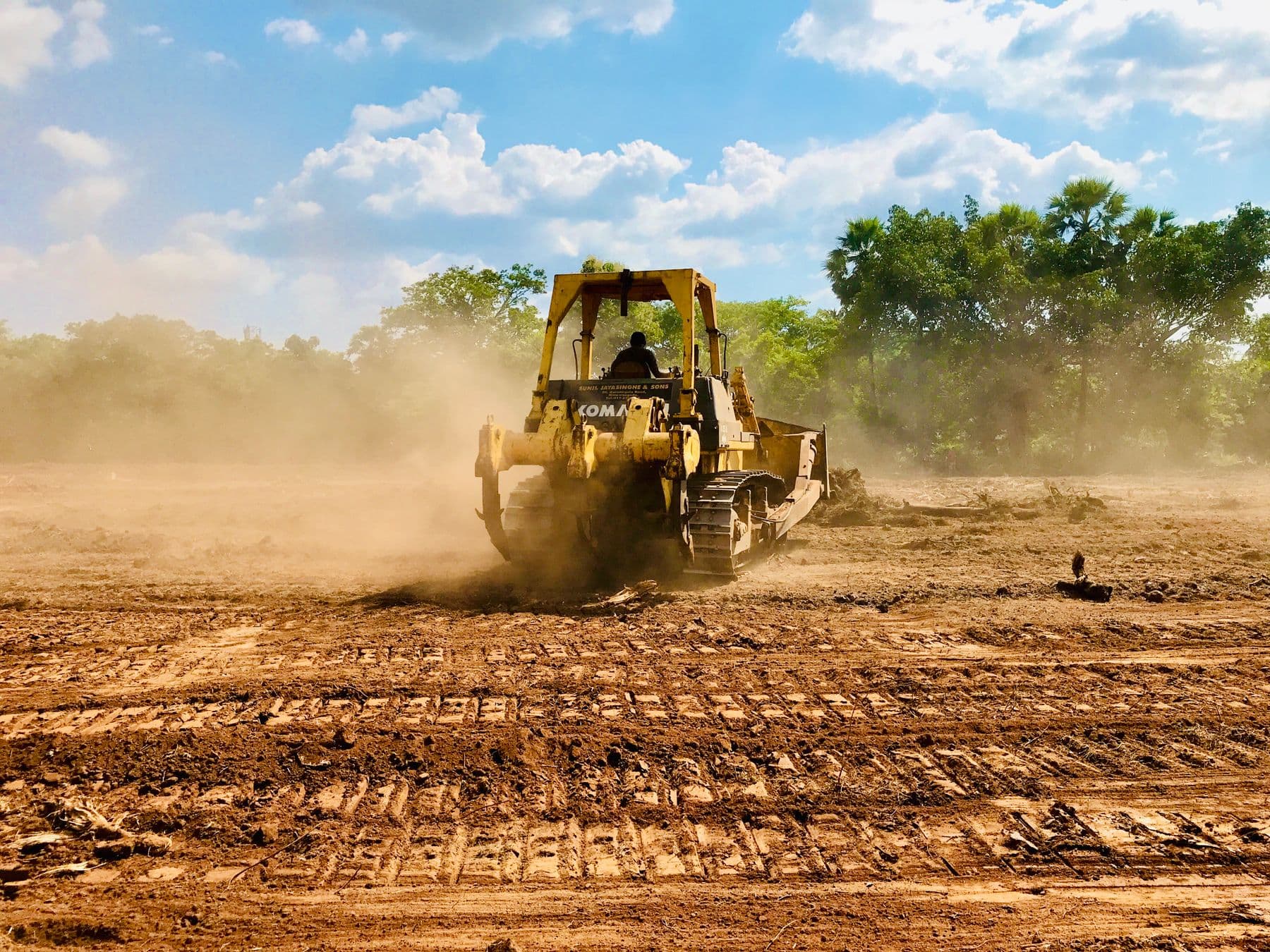 A yellow bulldozer raising dust as it grades a bare dirt lot, with trees beyond.