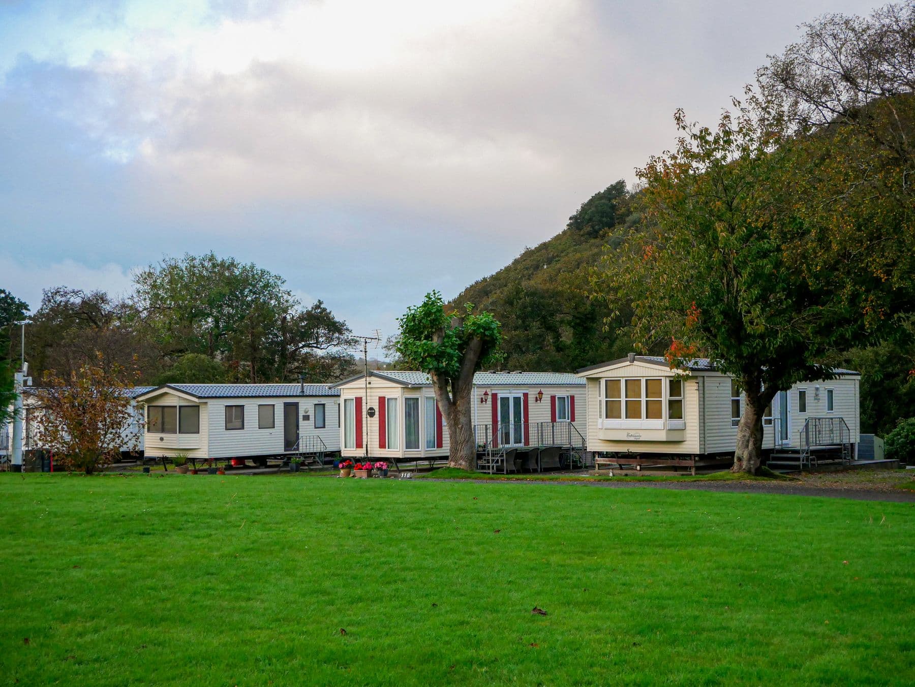 A row of mobile homes on a mown lawn, with trees and a hill behind them.