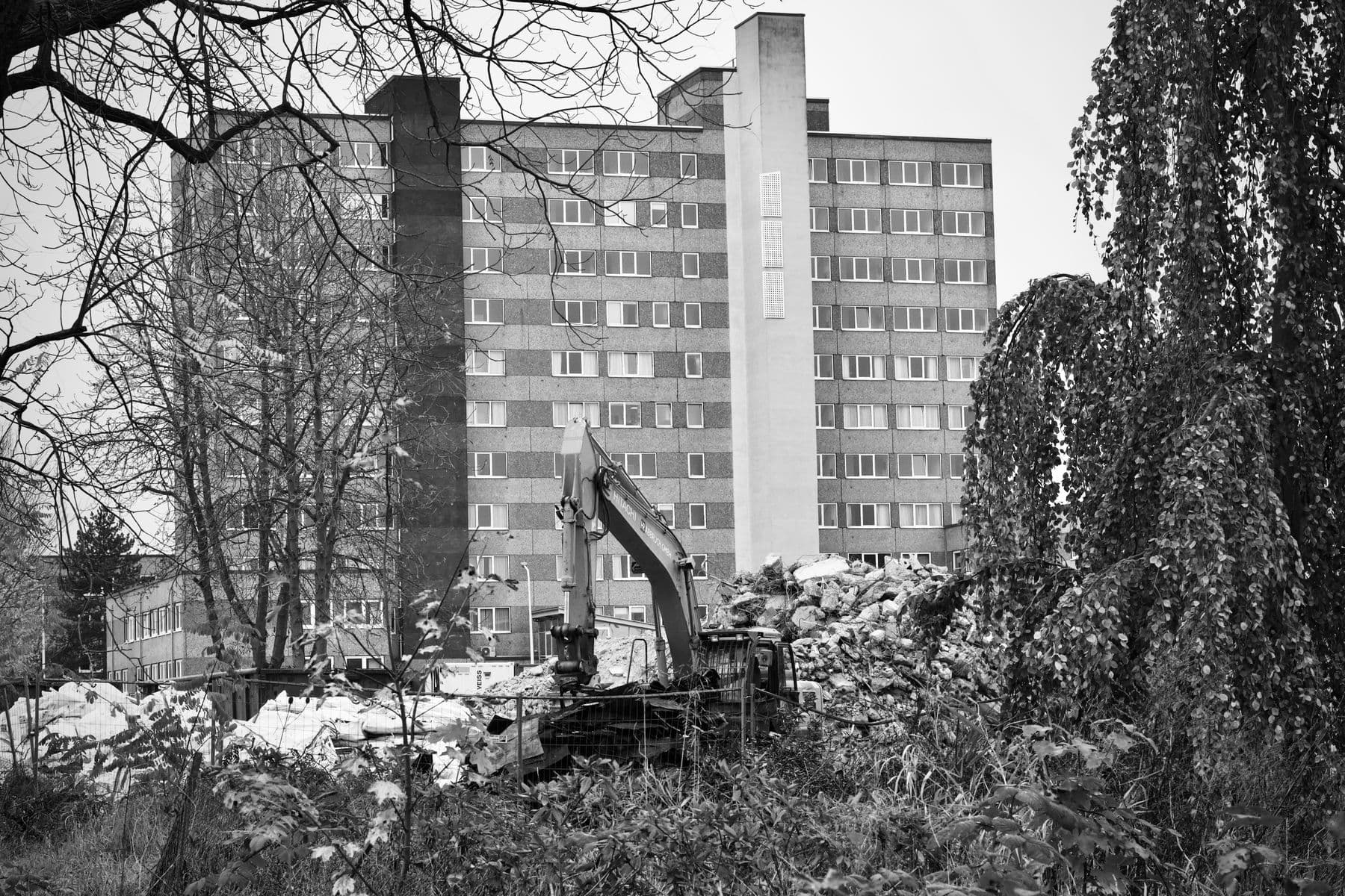 A black-and-white photo of an excavator working in rubble in front of a tall slab-block apartment building.